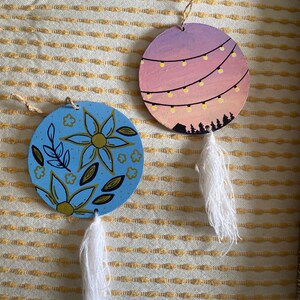 May include: Two round wooden ornaments with white tassels. One ornament features a blue background with gold floral designs. The other ornament features a pink and purple sunset with white string lights.