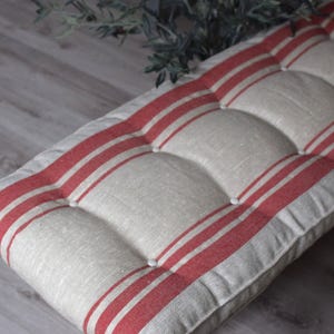 May include: A rectangular bench cushion featuring a natural beige fabric base with bold, vertical red stripes. The cushion is tufted with matching fabric-covered buttons, creating a series of raised, padded sections across the entire surface.