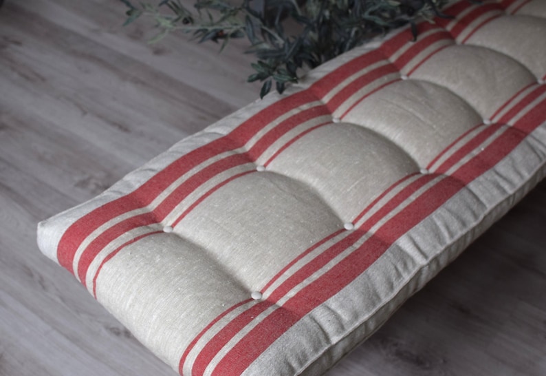 May include: A rectangular bench cushion featuring a natural beige fabric base with bold, vertical red stripes. The cushion is tufted with matching fabric-covered buttons, creating a series of raised, padded sections across the entire surface.