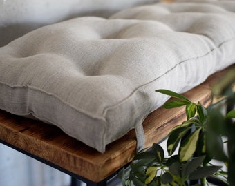 May include: A light gray linen cushion with button tufting sits on a wooden bench with a dark brown finish. The bench has a black metal frame and is partially obscured by a green leafy plant.