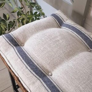 May include: A rectangular bench cushion made of natural beige linen fabric featuring two dark blue vertical stripes running along the length. The cushion is tufted with matching fabric-covered buttons and rests on a dark metal frame base.