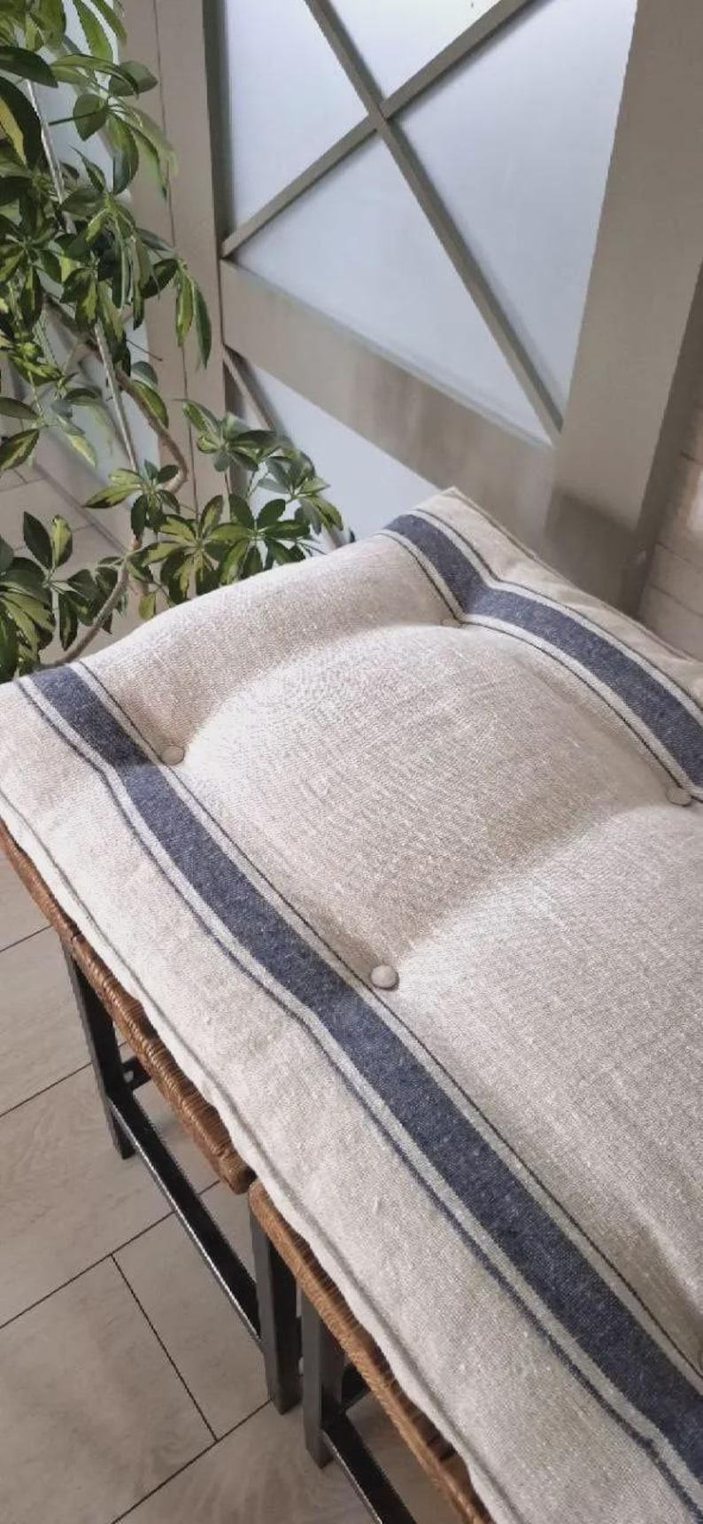 May include: A rectangular bench cushion made of natural beige linen fabric featuring two dark blue vertical stripes running along the length. The cushion is tufted with matching fabric-covered buttons and rests on a dark metal frame base.