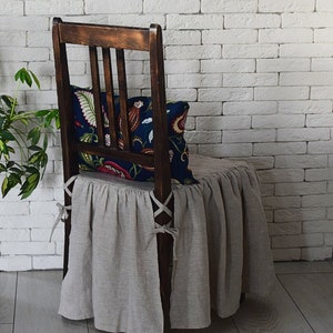 May include: A brown wooden chair with a linen chair cover and a floral patterned pillow. The chair is in front of a white brick wall.