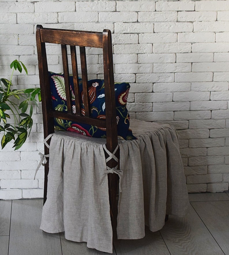 May include: A brown wooden chair with a linen chair cover and a floral patterned pillow. The chair is in front of a white brick wall.