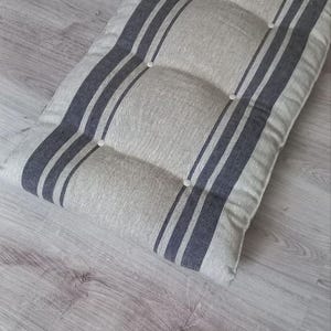 May include: A rectangular bench cushion featuring a light beige fabric base with dark gray vertical stripes. The cushion is tufted with small white buttons arranged in two rows along the length, creating a padded, textured surface appearance.