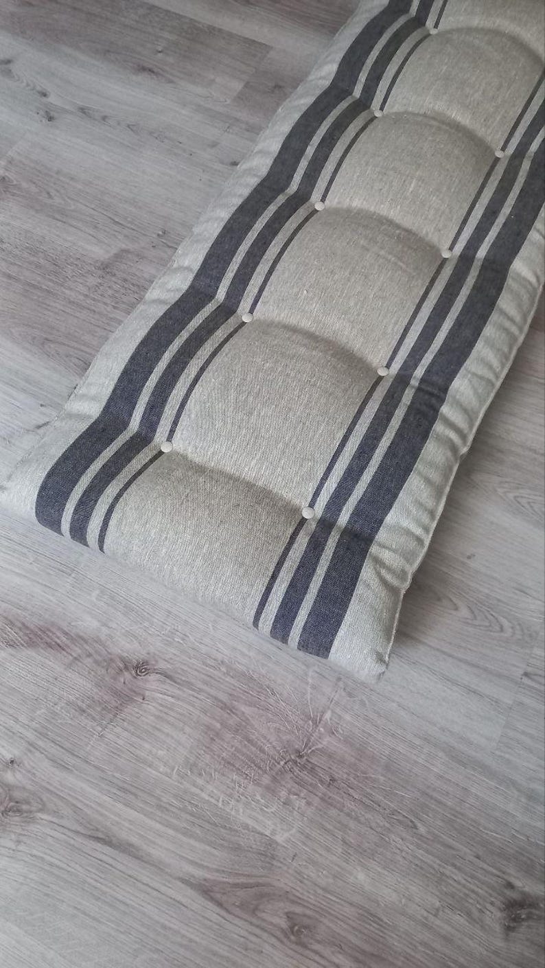 May include: A rectangular bench cushion featuring a light beige fabric base with dark gray vertical stripes. The cushion is tufted with small white buttons arranged in two rows along the length, creating a padded, textured surface appearance.