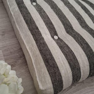 May include: A black and white striped floor cushion with button tufting. The cushion is made of a linen fabric and has a rectangular shape.