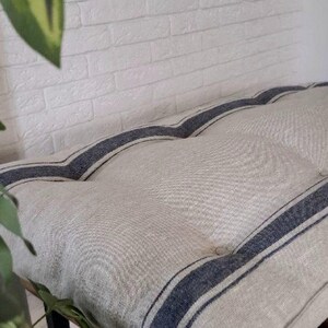 May include: A rectangular bench cushion featuring a natural beige linen-like fabric with two prominent dark blue vertical stripes running the length of the piece. The cushion is tufted with visible buttons and rests on a dark metal frame base.