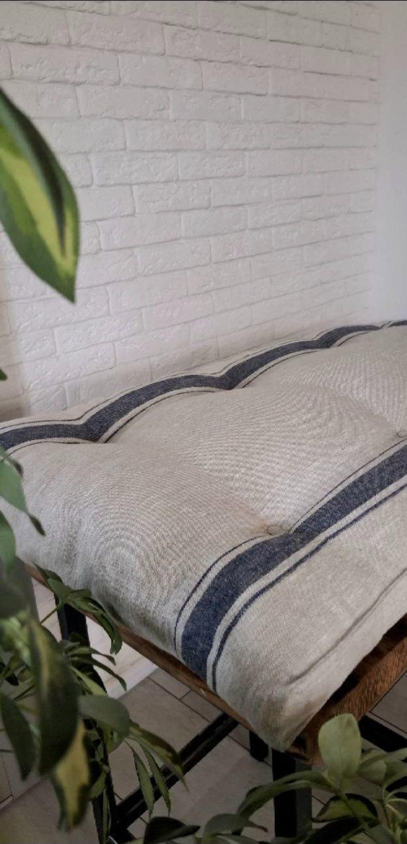 May include: A rectangular bench cushion featuring a natural beige linen-like fabric with two prominent dark blue vertical stripes running the length of the piece. The cushion is tufted with visible buttons and rests on a dark metal frame base.