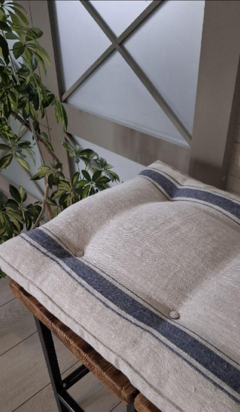 May include: A square seat cushion made of natural beige fabric featuring two dark blue vertical stripes and two matching fabric-covered buttons. The cushion has a tufted design and is positioned on a wooden surface with a visible woven texture.