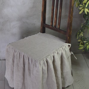 May include: A beige linen chair cover with a ruffled edge and a tie at the back. The cover is on a dark wooden chair.