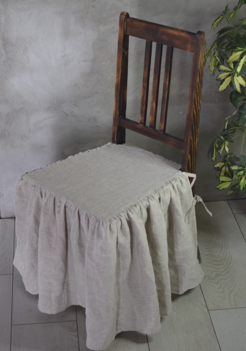 May include: A beige linen chair cover with a ruffled edge and a tie at the back. The cover is on a dark wooden chair.
