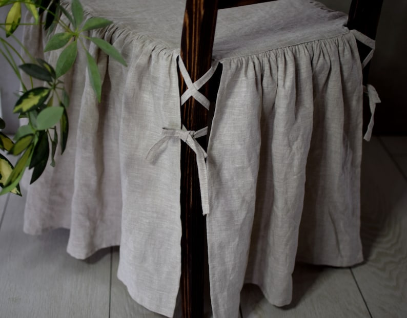May include: A beige linen chair cover with a ruffled bottom and ties on the sides. The cover is tied around the back legs of the chair.