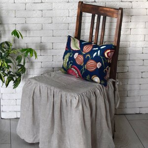May include: A beige linen chair cover with a ruffled edge, covering a wooden chair with a floral patterned pillow. The chair is in front of a white brick wall.
