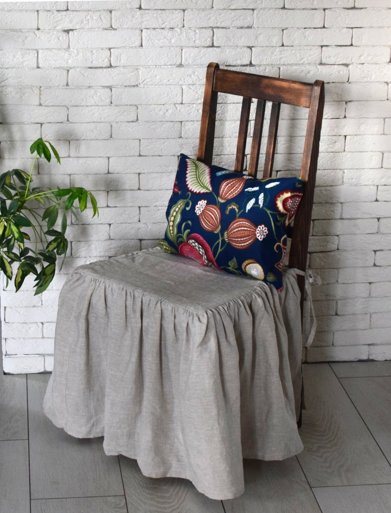 May include: A beige linen chair cover with a ruffled edge, covering a wooden chair with a floral patterned pillow. The chair is in front of a white brick wall.