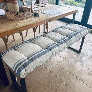 May include: A rectangular bench with a padded cushion. The cushion is made of a light-colored fabric with blue and white stripes. The bench has a dark metal frame and is placed on a tiled floor.