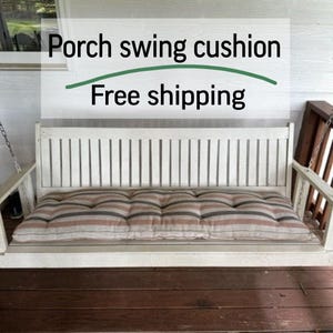Può includere: Un'altalena da veranda in legno bianco con un cuscino a righe. Il cuscino presenta strisce marroni, grigie e crema. Il testo "Porch swing cushion" è visibile sopra l'altalena.