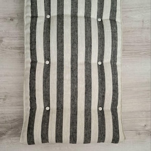 May include: A rectangular floor cushion featuring a vertical striped pattern in charcoal gray and off-white. The cushion is tufted with eight small, round, white buttons arranged in two columns, creating a textured, padded surface appearance.