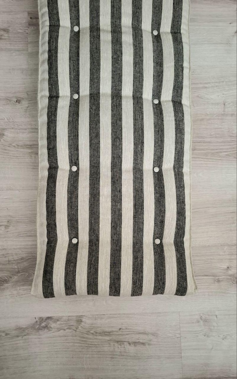 May include: A rectangular floor cushion featuring a vertical striped pattern in charcoal gray and off-white. The cushion is tufted with eight small, round, white buttons arranged in two columns, creating a textured, padded surface appearance.