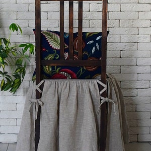 May include: A wooden chair with a beige linen chair skirt and a floral patterned pillow. The chair is in front of a white brick wall.