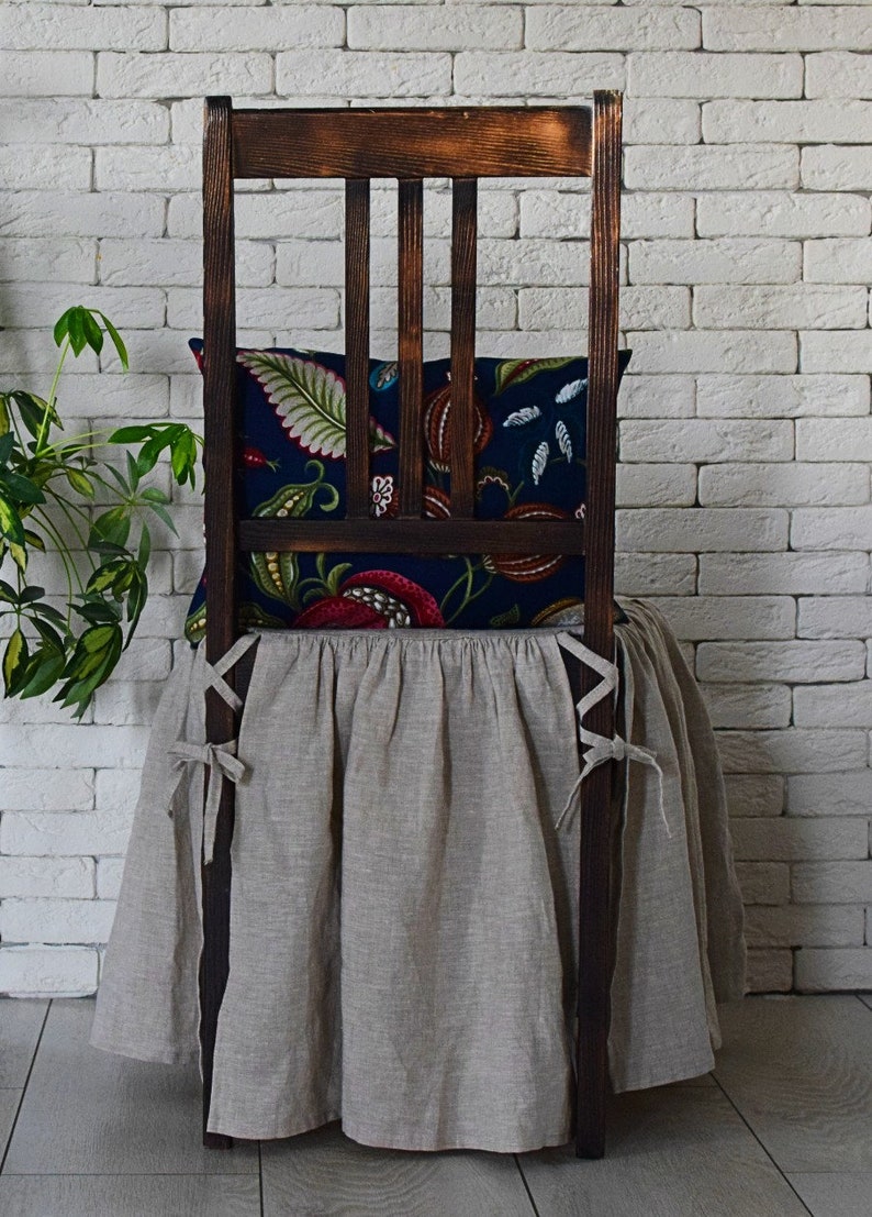 May include: A wooden chair with a beige linen chair skirt and a floral patterned pillow. The chair is in front of a white brick wall.