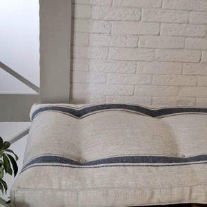 May include: A rectangular bench cushion made of natural beige fabric featuring two horizontal dark blue stripes that curve in a wave pattern across the surface. The cushion has a tufted design with visible indentations and a structured edge.