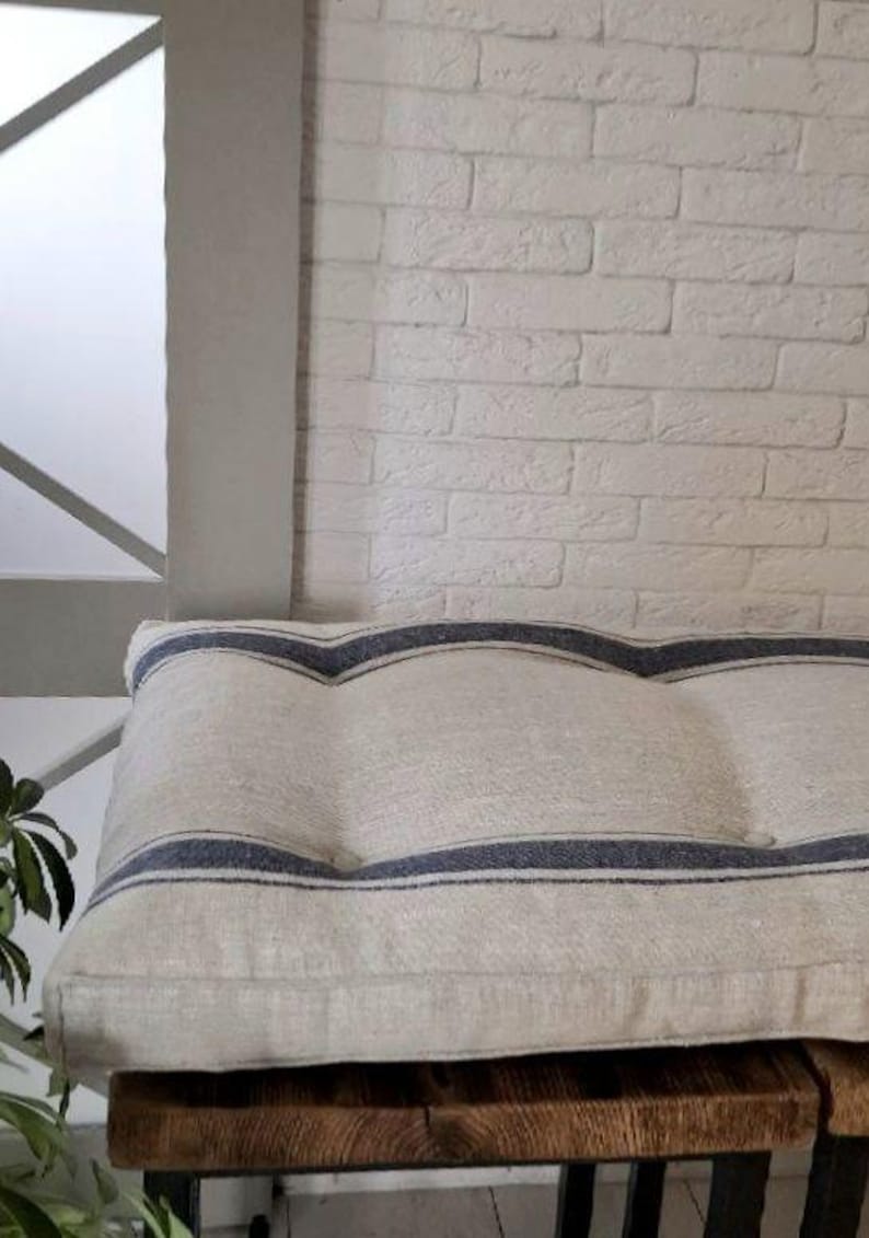 May include: A rectangular bench cushion made of natural beige fabric featuring two horizontal dark blue stripes that curve in a wave pattern across the surface. The cushion has a tufted design with visible indentations and a structured edge.