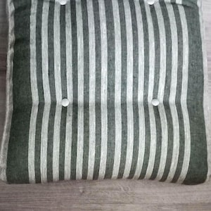 May include: A rectangular seat cushion featuring a vertical striped pattern in dark green and off-white. The cushion is tufted with four circular white buttons arranged in two rows, creating a textured surface with distinct indentations.