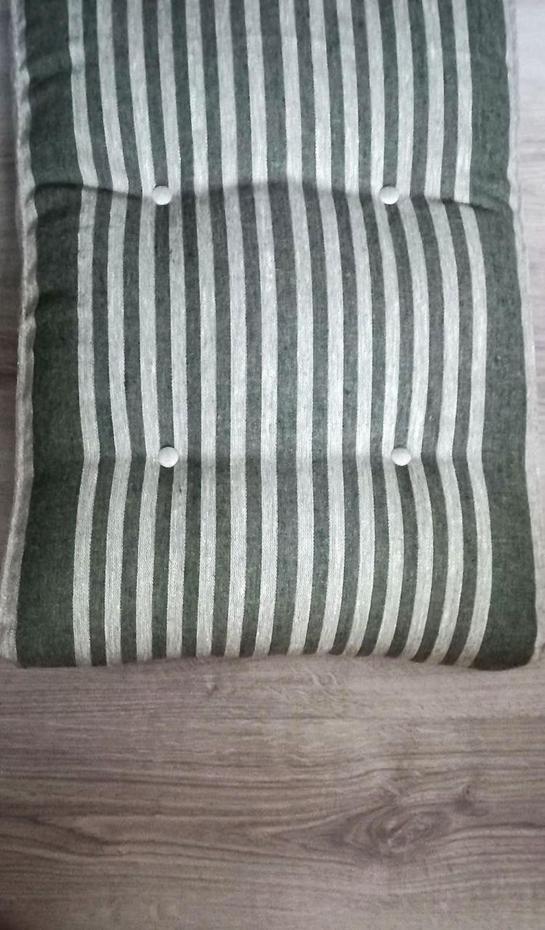 May include: A rectangular seat cushion featuring a vertical striped pattern in dark green and off-white. The cushion is tufted with four circular white buttons arranged in two rows, creating a textured surface with distinct indentations.