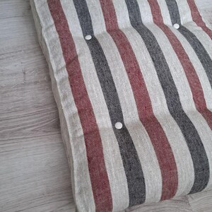 May include: A rectangular floor cushion featuring a vertical striped pattern in shades of dark gray, muted red, and off-white. The cushion is tufted with small, round, white buttons arranged in a grid pattern across the textured fabric surface.