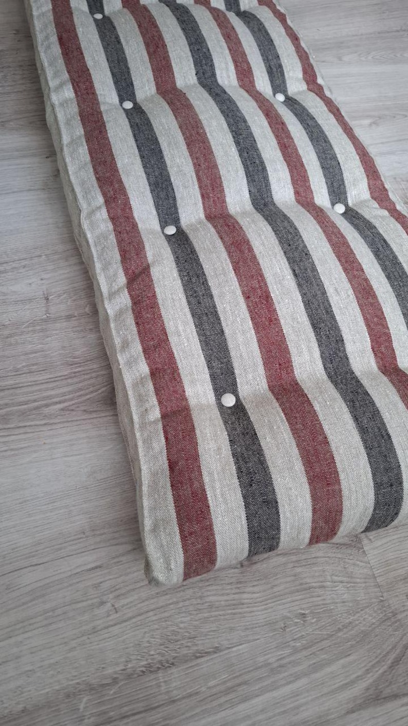 May include: A rectangular floor cushion featuring a vertical striped pattern in shades of dark gray, muted red, and off-white. The cushion is tufted with small, round, white buttons arranged in a grid pattern across the textured fabric surface.