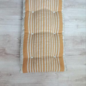 May include: A rectangular floor cushion featuring a vertical striped pattern in mustard yellow and off-white. The cushion is tufted with six small white circular buttons arranged in two parallel rows, creating a soft, padded, and textured surface.
