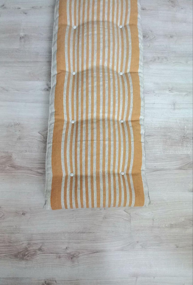 May include: A rectangular floor cushion featuring a vertical striped pattern in mustard yellow and off-white. The cushion is tufted with six small white circular buttons arranged in two parallel rows, creating a soft, padded, and textured surface.