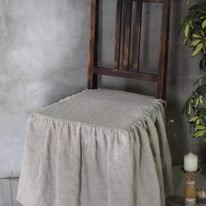 May include: A beige linen chair cover with a ruffled edge, draped over a dark wooden chair with a slatted back.