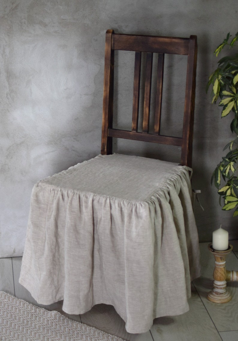 May include: A beige linen chair cover with a ruffled edge, draped over a dark wooden chair with a slatted back.