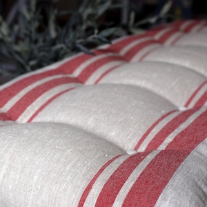 May include: A rectangular bench cushion featuring a tufted design with a series of deep indentations. The fabric is a natural beige color accented with bold, vertical red stripes running the length of the cushion, creating a classic striped pattern.