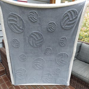 May include: A gray blanket with an embossed volleyball pattern. The blanket has a white, fluffy border. The volleyballs are in various sizes and are evenly spaced across the blanket's surface. The blanket is draped over a chair.