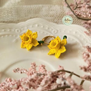 Op de afbeelding: Een paar gehaakte narcissen oorbellen met gouden hoepels. De oorbellen liggen op een wit bord met een roze bloem op de achtergrond.