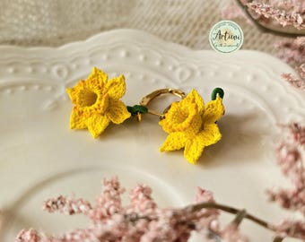 May include: A pair of crocheted daffodil earrings with gold hoops. The earrings are on a white plate with a pink flower in the background.