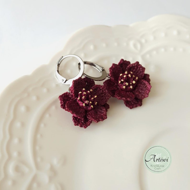 May include: A pair of burgundy crocheted flower earrings with gold accents hanging from silver hoop earrings.