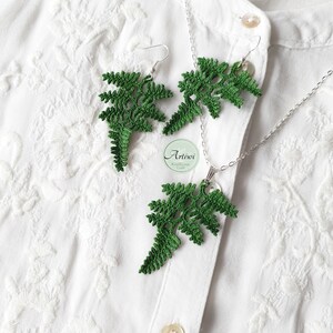 May include: A set of green crocheted fern earrings and a matching necklace. The earrings are dangling and the necklace has a pendant in the shape of a fern. The jewellery is displayed on a white shirt.