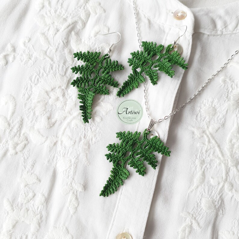 May include: A set of green crocheted fern earrings and a matching necklace. The earrings are dangling and the necklace has a pendant in the shape of a fern. The jewellery is displayed on a white shirt.