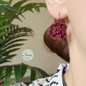May include: Burgundy crocheted flower earrings with gold accents. The earrings are hanging from a person's ear.