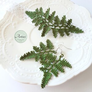 May include: A pair of green crocheted fern earrings with silver hooks. The earrings are displayed on a white plate with a lace doily.