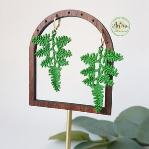 May include: Green crocheted fern earrings hanging from gold hoops on a wooden earring display stand. The stand has a rounded arch top and is on a gold metal rod.