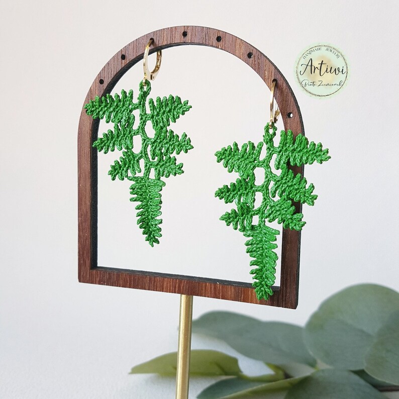 May include: Green crocheted fern earrings hanging from gold hoops on a wooden earring display stand. The stand has a rounded arch top and is on a gold metal rod.