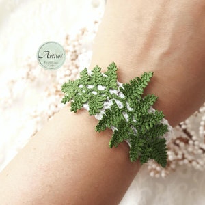 May include: A white crocheted bracelet with a green crocheted fern design. The bracelet is on a person's wrist. The text "Artivoi Knots&Loop Craft" is on a white circle.