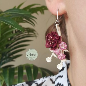 May include: Burgundy and pink crocheted flower earrings with a silver hook. The earrings are hanging from a person's ear.