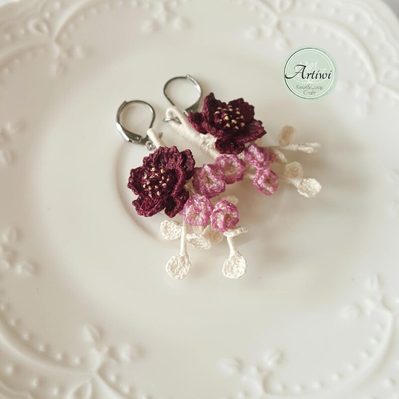 May include: A pair of crocheted flower earrings with burgundy and pink flowers and white stems. The earrings are on a white plate with a decorative edge.