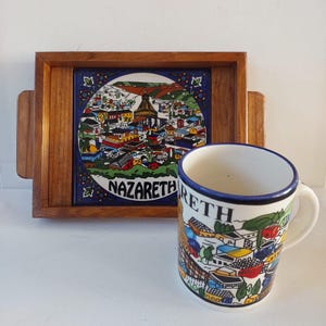 May include: A wooden tray with a blue and white ceramic tile depicting a colorful cityscape with the word "Nazareth" on it. A white ceramic mug with a blue rim and a similar cityscape design is next to the tray.